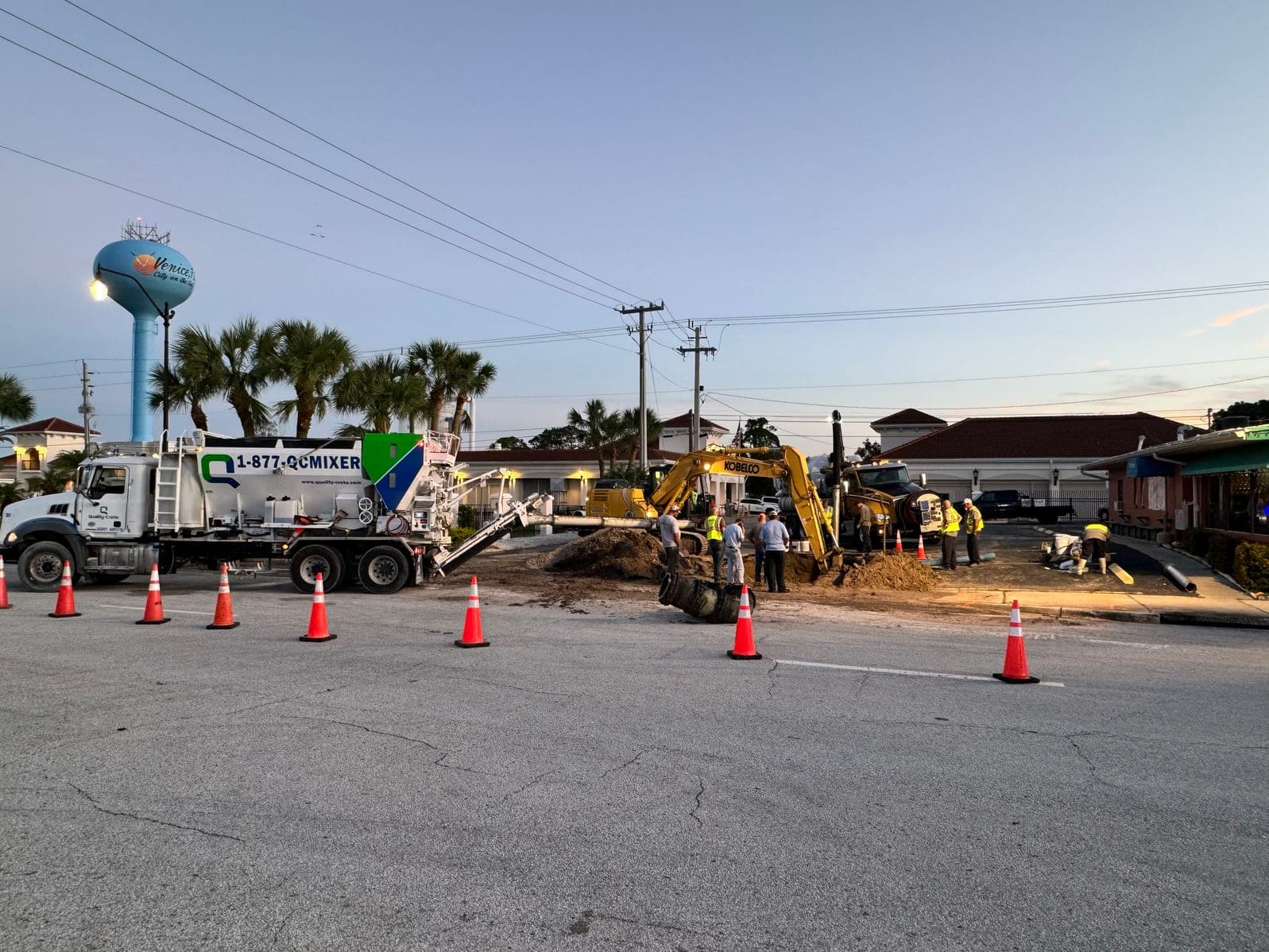 Quality-Crete volumetric mixer truck beside an excavator and crew at a roadside utility repair at dusk