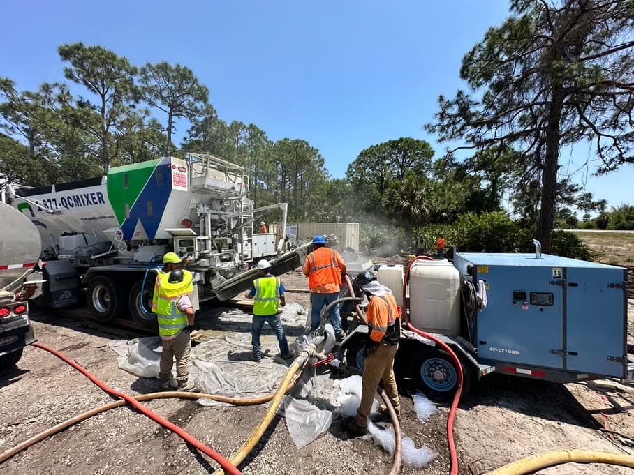 Crew connecting hoses to a Quality-Crete volumetric mixer and pump trailer on a wooded jobsite